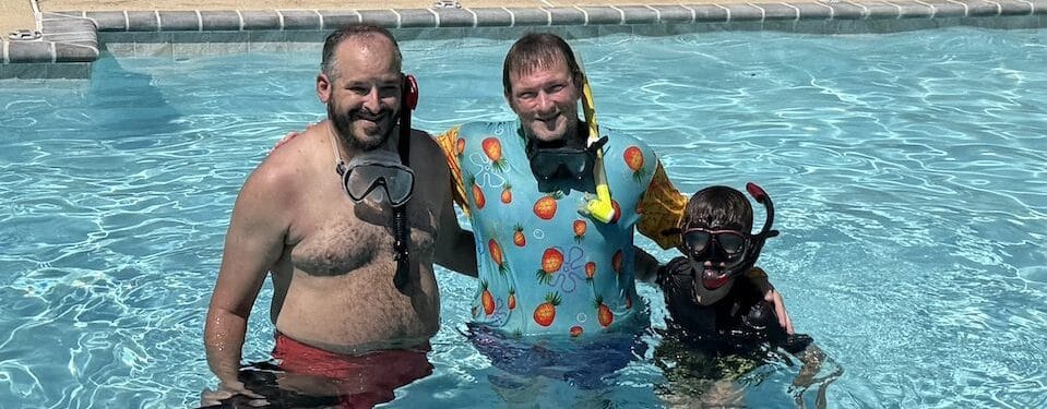 Three scuba divers smiling in a pool after training, one wearing a colorful shirt.