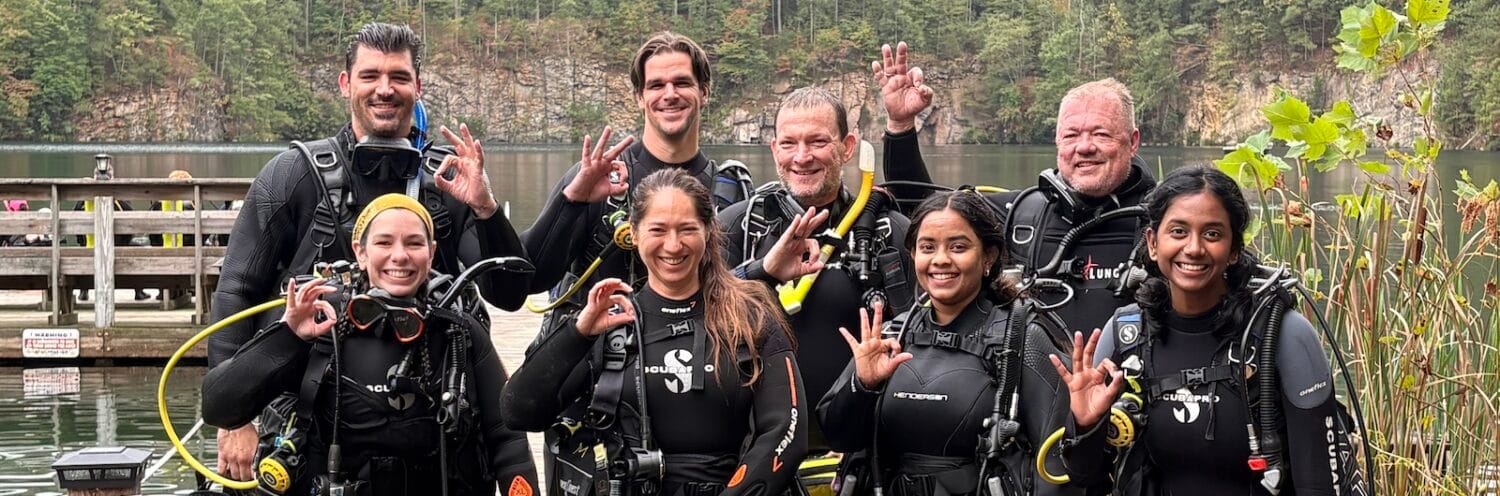 Group photo of eight people (seven students + instructor) celebrating Open Water Diver certification at Lake Phoenix.