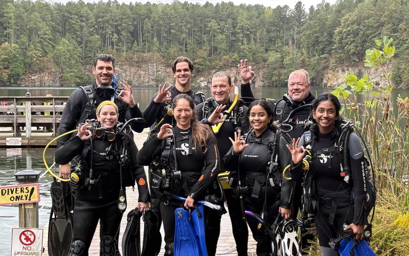 Group photo of eight people (seven students + instructor) celebrating Open Water Diver certification at Lake Phoenix.