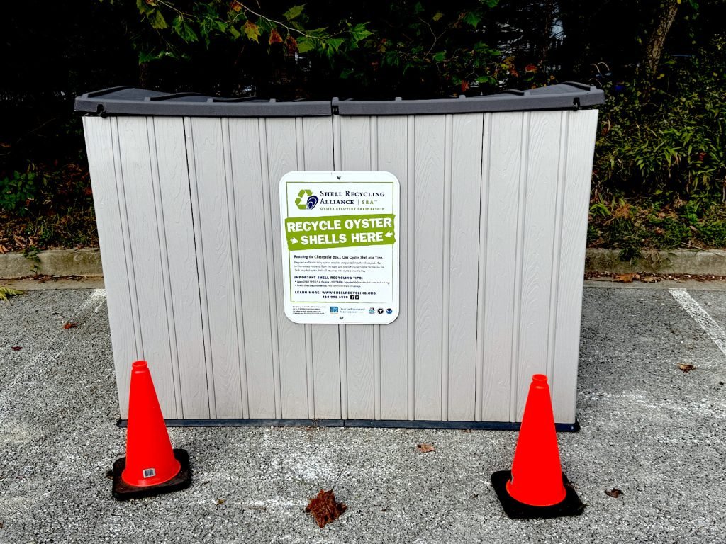 Gray recycling bin with a green sign reading “Recycle Oyster Shells Here.”