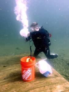 Diver kneeling underwater attaching a line to an orange Home Depot bucket and white jug during the Zombie Apocalypse Diver course.