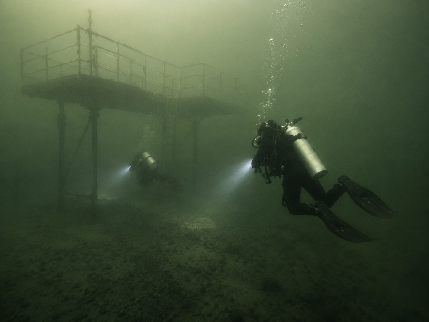 Two scuba divers use lights near an underwater training platform in low-visibility green water.